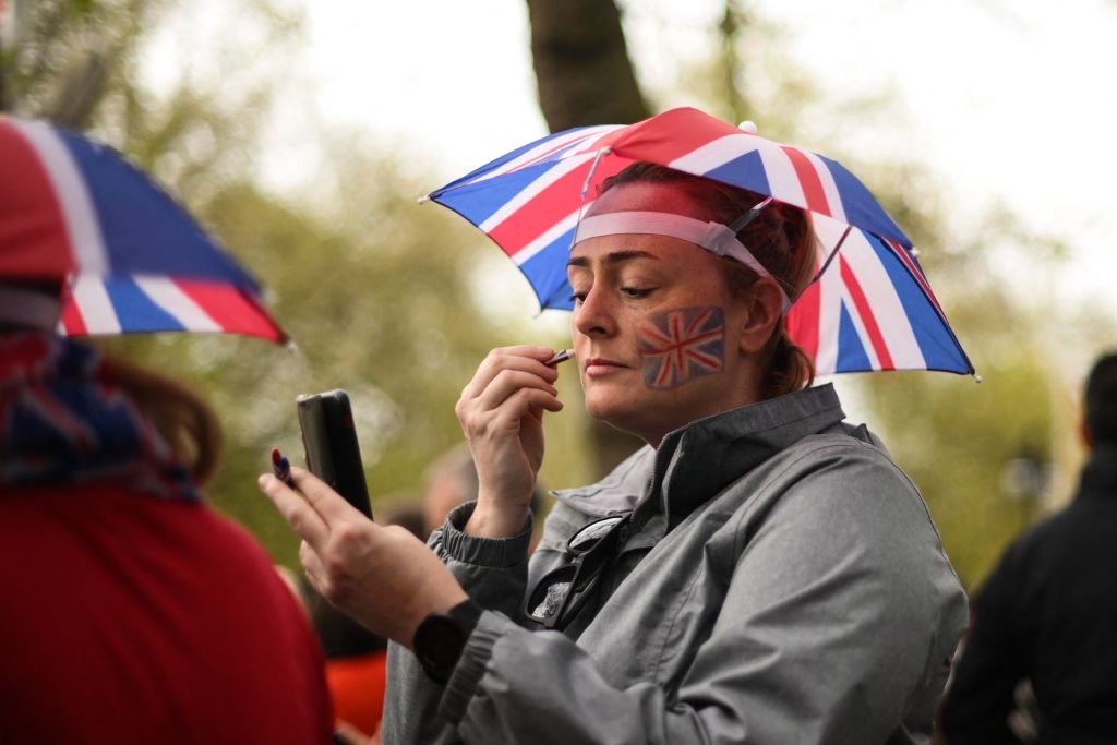 A woman applies facepaint while wearing a Union Jack umbrella hat on The Mall