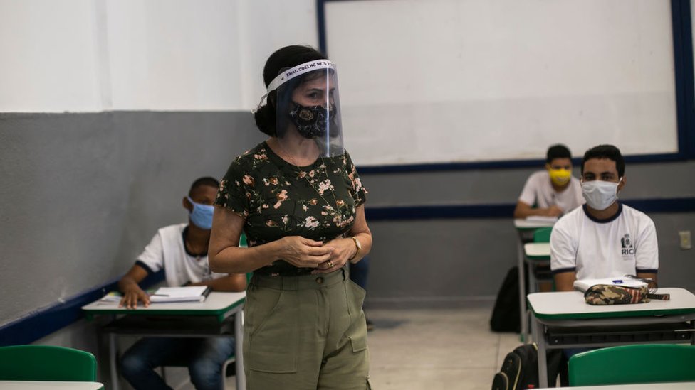 Professora em pé com máscara e protetor facial. Alunos, que estão sentados, usam mácaras e uniforme.