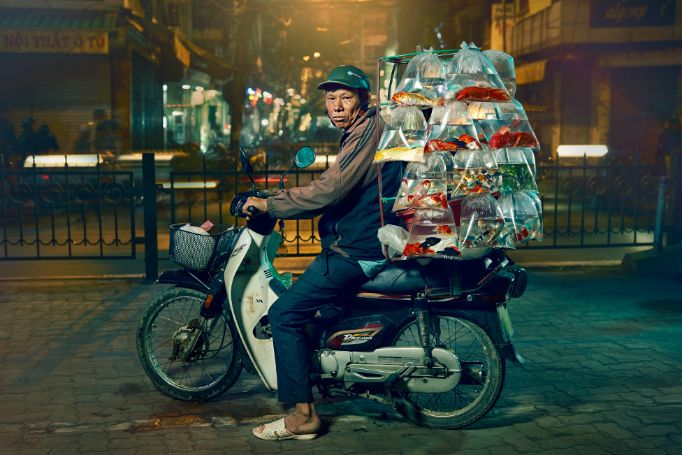 Мужчина сидит на мотоцикле с множеством разноцветных рыбок в пластиковых пакетах, рядами висящих на задней части мотоцикла