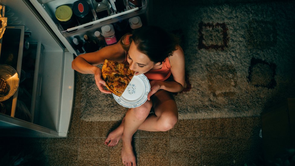 Mujer comiendo con la puerta del frigorífico abierta.