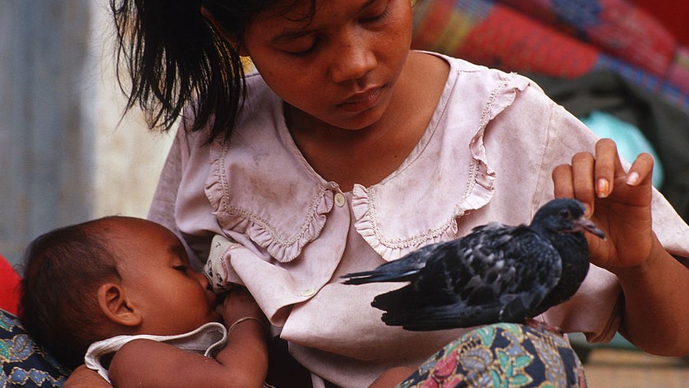 A young Cambodian mother feeding her baby and petting a pigeon