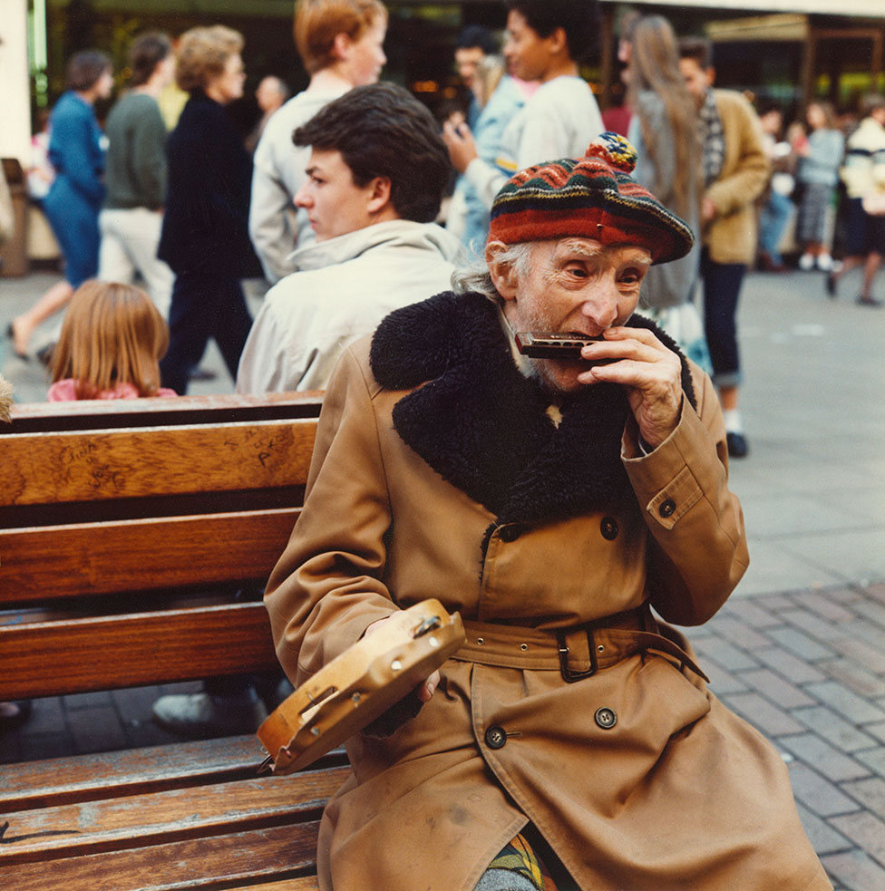 Пожилой мужчина сидит на уличной скамейке и играет на губной гармошке