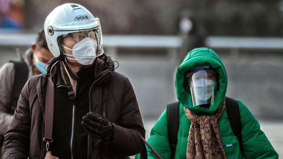 Um homem usando capacete e máscara facial n95 caminha pelas ruas e uma mulher usando máscara e protetor facial atrás dele