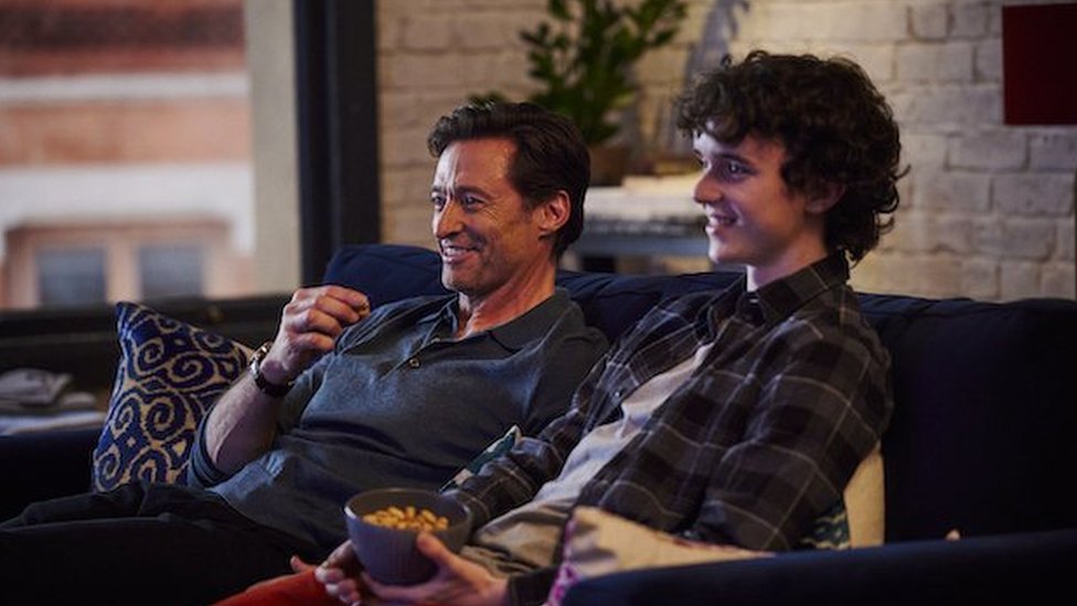 Hugh Jackman e Zen McGrath sentados no sofá rindo durante as filmagens de 'Um Filho'