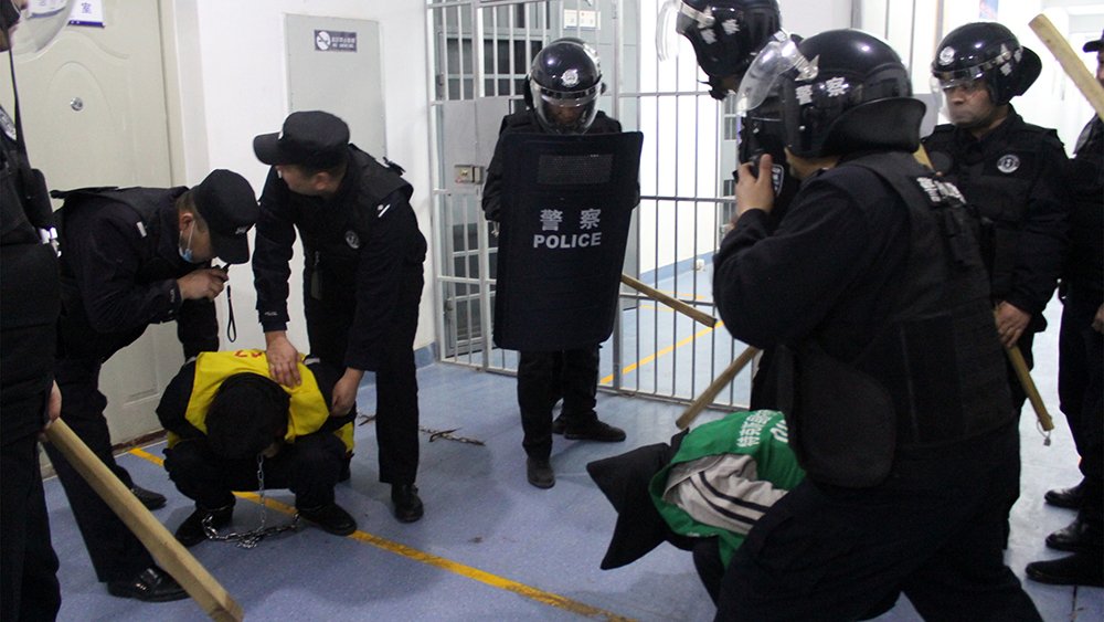 A detainee squats, head down, while surrounded by officers with batons