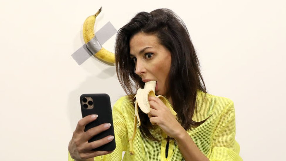 Una mujer comiendo una banana mientras se toma un selfie con la banana expuesta.