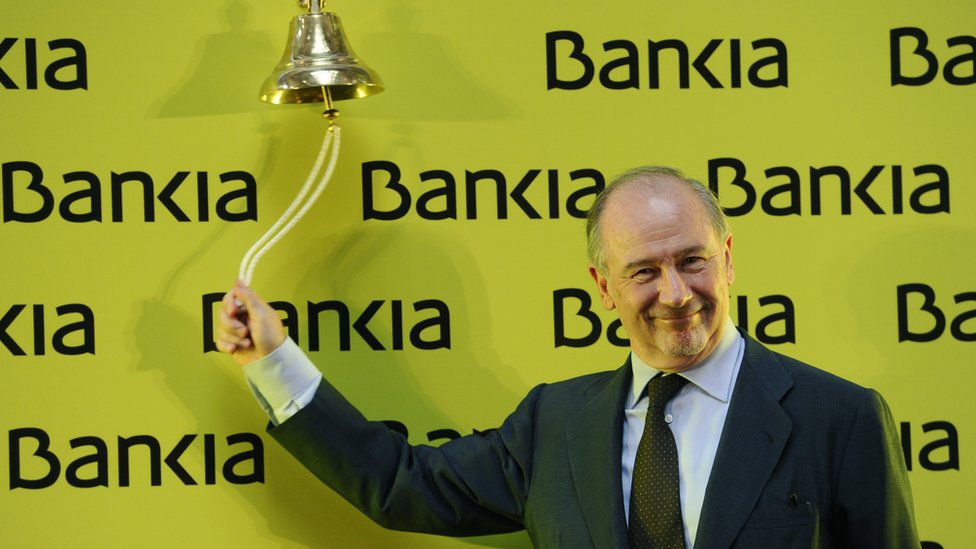 Родриго Рато, улыбаясь, стоит напротив желтого плаката с надписью «Bankia!» И звонит в звонок на Мадридской фондовой бирже 20 июля 2011 года, чтобы отметить начало листинга Bankia