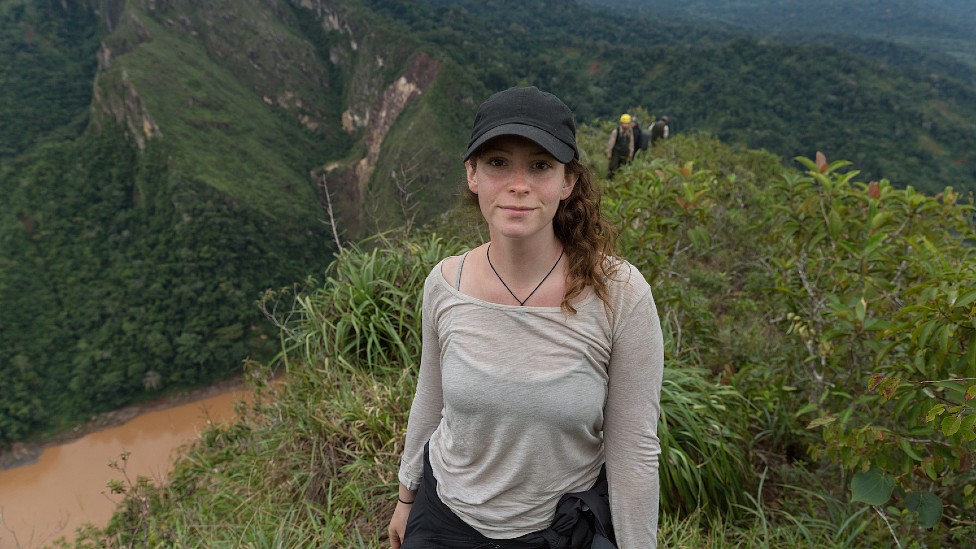 A diretora Elizabeth Unger, uma mulher branca jovem, em trilha em meio a montanhas cobertas por floresta