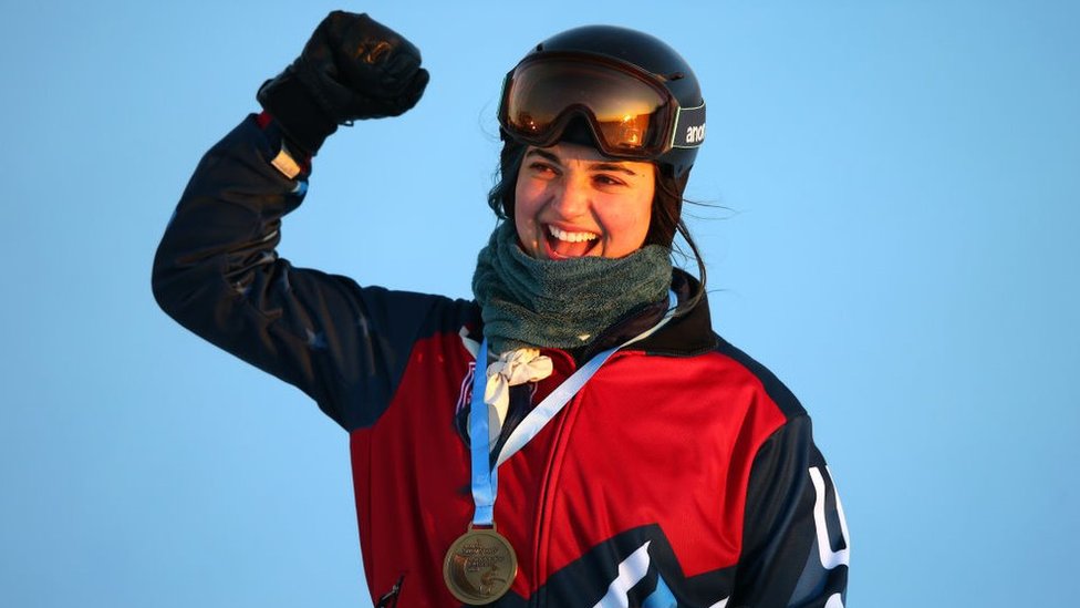 Huckaby punching the air, wearing a snowboarding helmet and goggles with a gold medal around her neck
