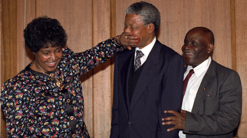 Винни Мандела игриво заставляет Нельсона Манделу замолчать, когда они покидают лондонскую пресс-конференцию в 1990 году