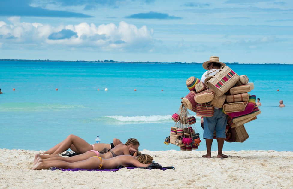 Hombre vendiendo artesanía en la playa mientras unos turistas descansan.