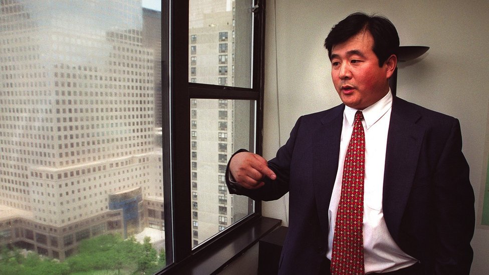Li Hong Zhi stands gesticulating in a business suit in an office overlooking other large buildings