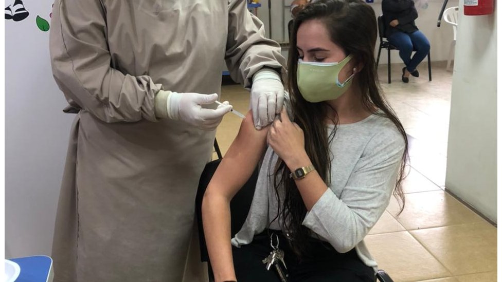 Daniela Muratório está sentada enquanto uma enfermeira aplica vacina contra a covid-19