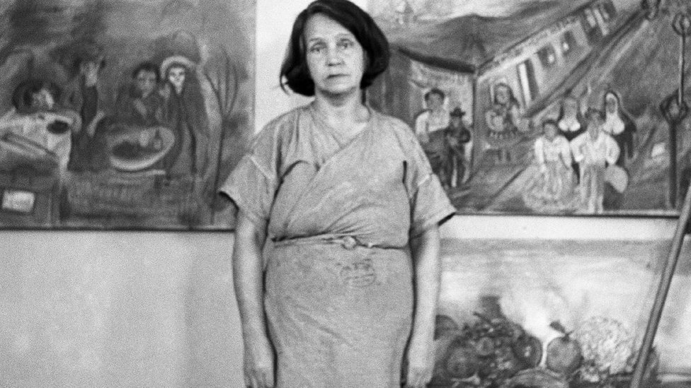 Fotografía en blanco y negro que muestra a una mujer con bata de laboratorio delante de varios cuadros