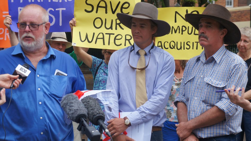 Трое фермеров на акции протеста с плакатом позади них: «Спасите нашу воду»