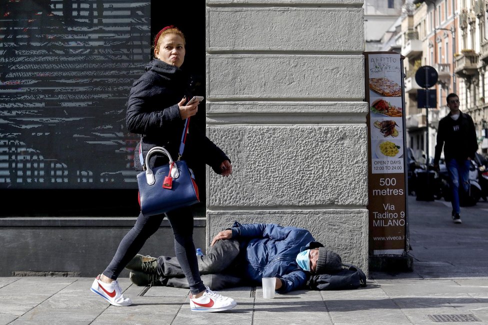 A woman walks by a homeless person who is wearing a face mask