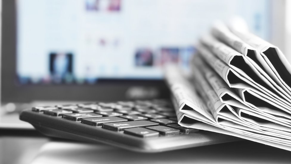Стопка сложенных газет сидит на клавиатуре перед монитором, на котором виден размытый новостной сайт