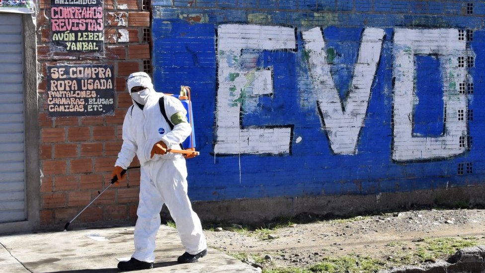 Trabajador de limpieza desinfectando una calle delante de una pinta de Evo Morales.