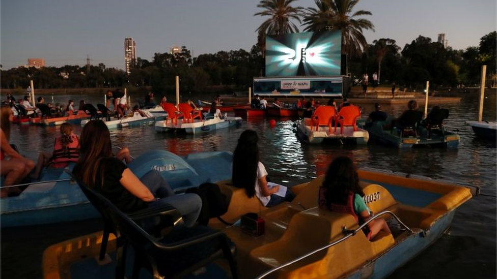 Grupos em embarcações no lago, de tarde, com tela de cinema ao fundo