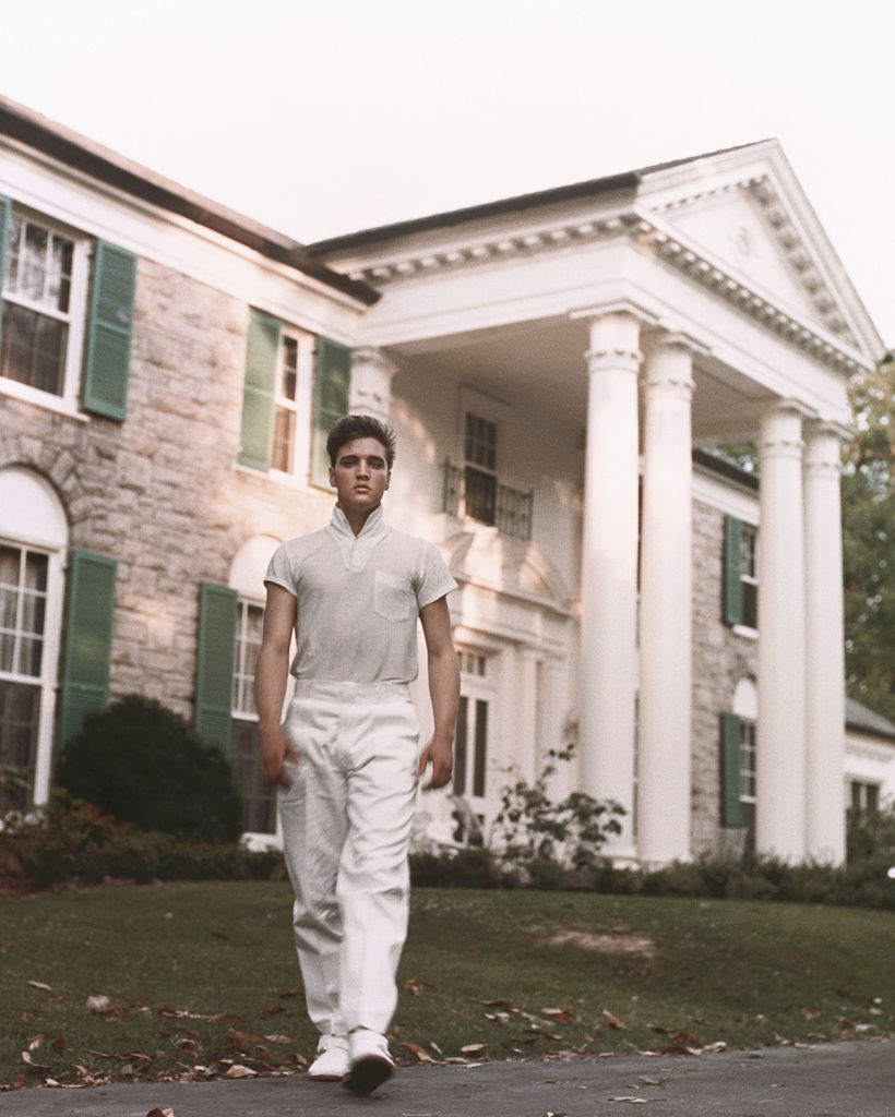 Elvis Presley, jovem, magro e vestido de branco, em frente à sua casa, que lhe custou 100 mil dólares em 1957
