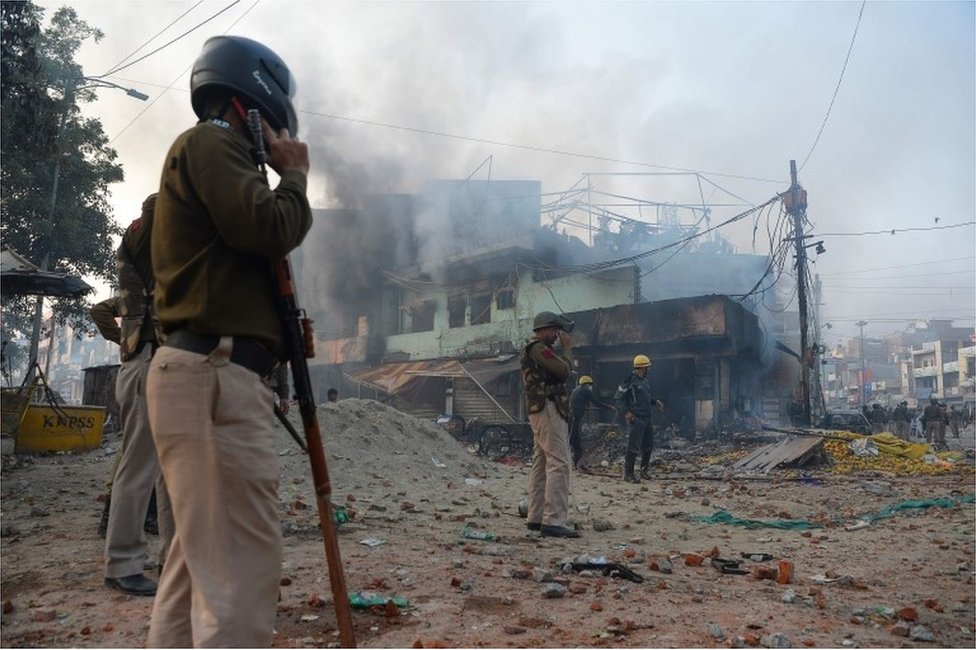 Полицейские стоят вдоль дороги, усыпанной камнями, в то время как клубы дыма из зданий после столкновений между сторонниками и противниками нового закона о гражданстве в районе Бхаджанпура в Нью-Дели 24 февраля 2020 г.