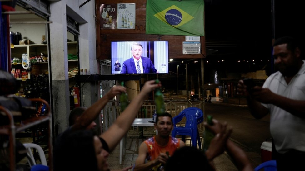 Bolsonaro na tela TV em bar, com pessoas conversando a frente