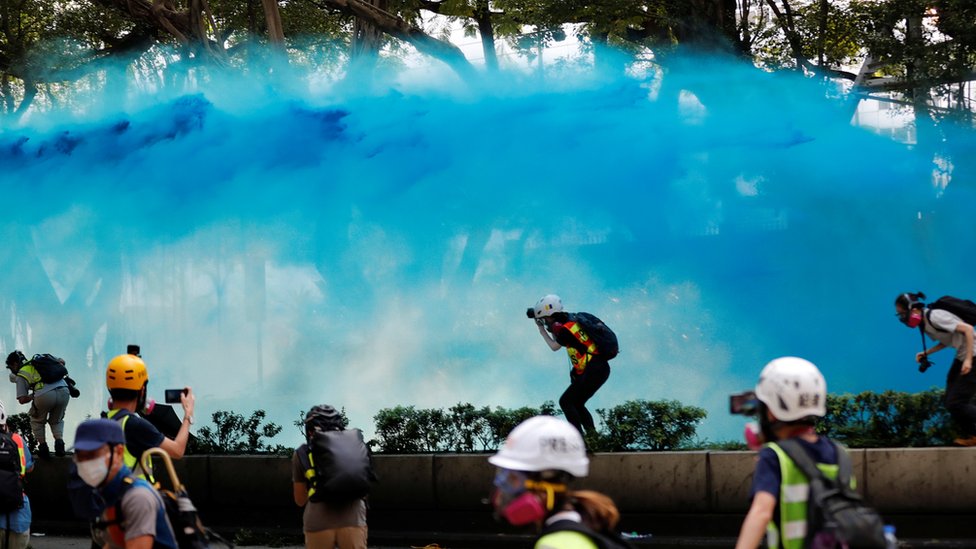 Волна сюрреалистической голубой воды проносится по воздуху, журналисты и протестующие ныряют, чтобы избежать попадания краски в открытое пространство