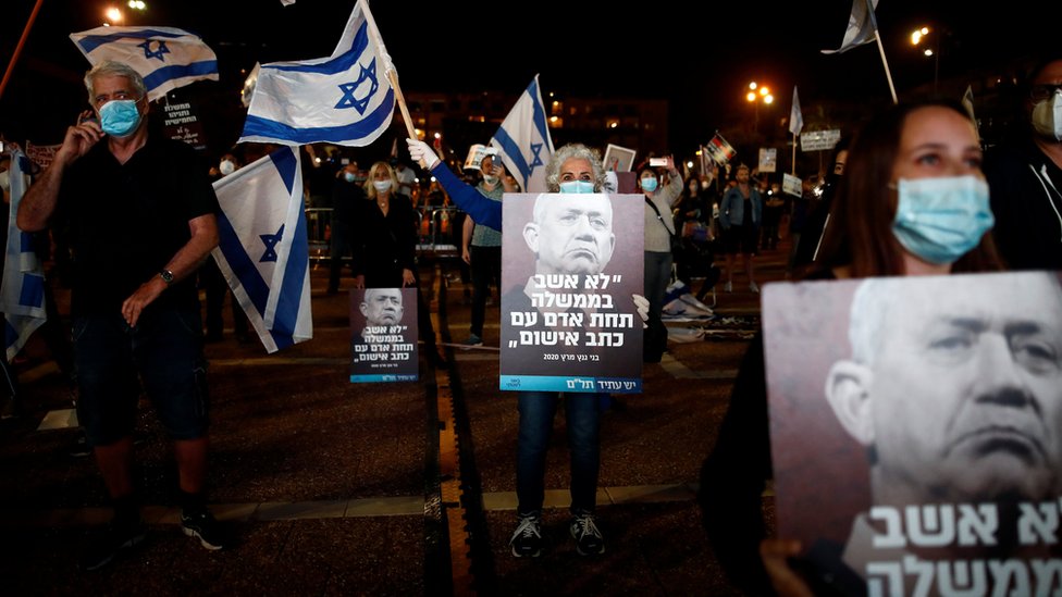 Люди держат плакаты с изображением Бенни Ганца и цитируют его слова: «Я не буду сидеть в правительстве под руководством человека, которому грозит уголовное обвинение» на протесте в Тель-Авиве 19 апреля 2020 года