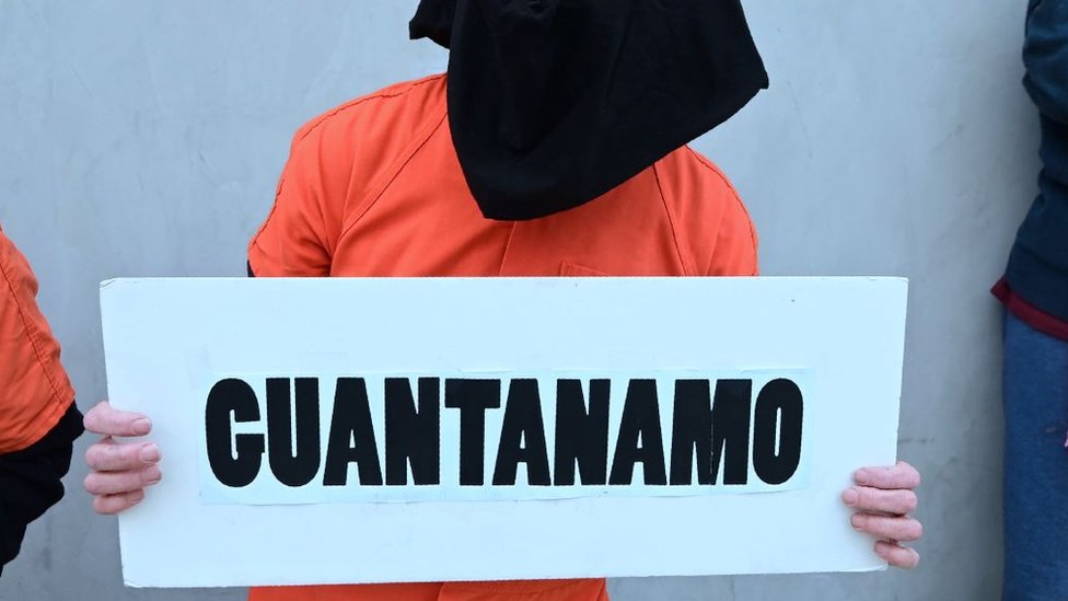 Homem com capuz segura cartaz escrito Guantánamo