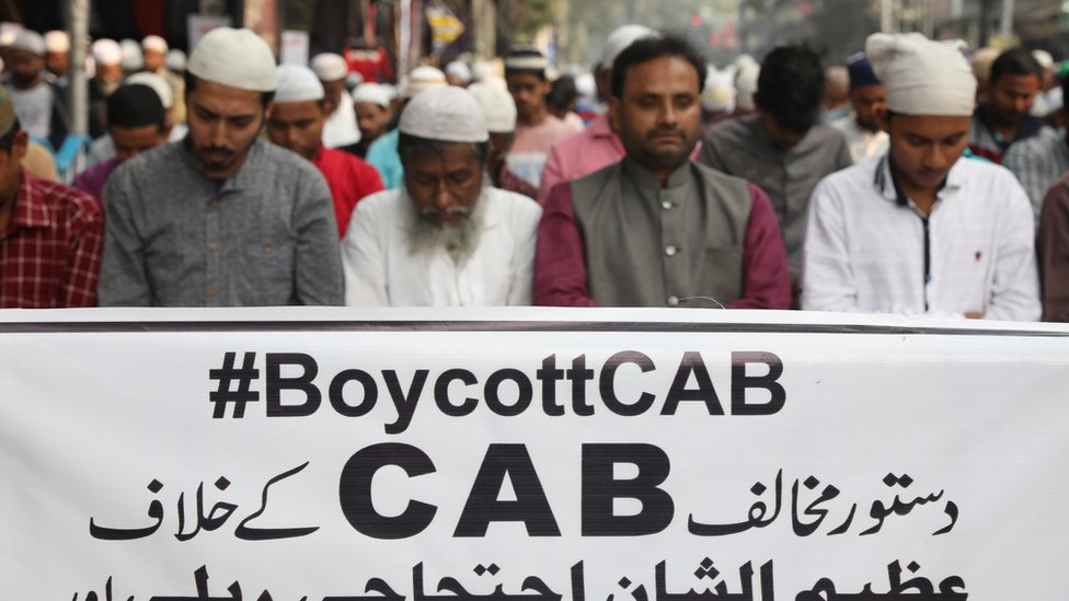 Протестующие совершают пятничную молитву перед транспарантом с надписью #BoycottCAB