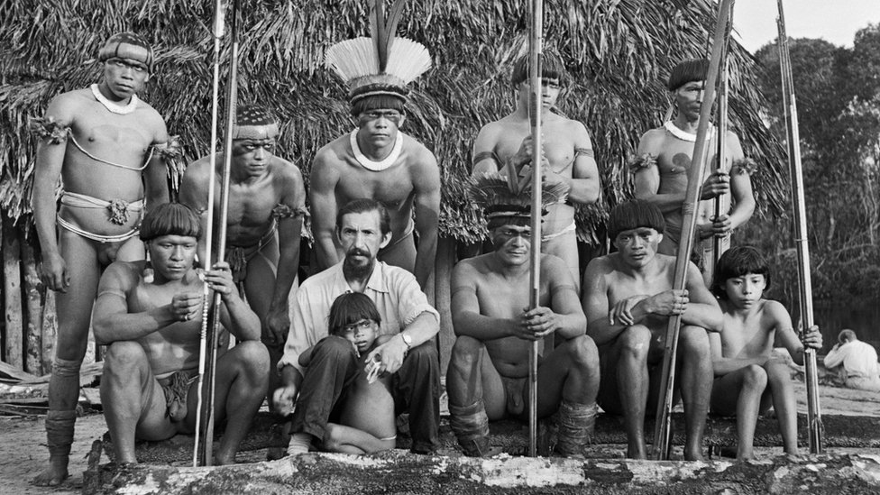 Fotografia em preto e branco mostra 9 homens do povo Yawalapiti, um homem branco e uma criança; parte do grupo está sentada