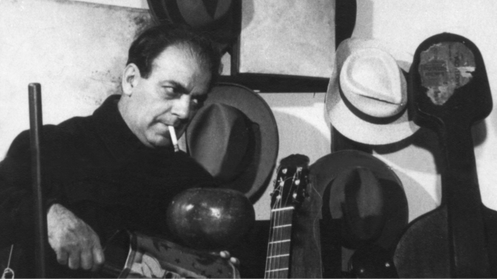 Fotografia em preto e branco do músico Heitor Villa-Lobos ao lado de uma coleção de chapéus e instrumentos musicais