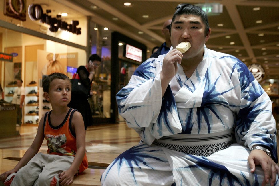 Японский сумоист Секиваке Котомицуки ест мороженое, пока маленький мальчик наблюдает за ним во время посещения торгового центра Тель-Авива
