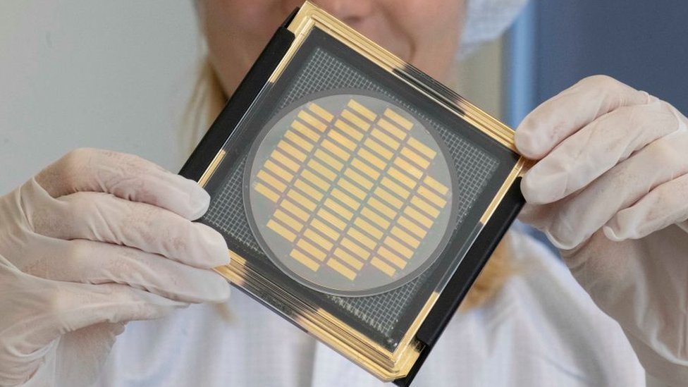 A scientist in "clean room" gear holds up a large computer chip wafer the size of a fist