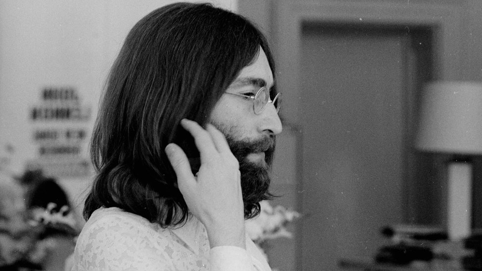Fotografia em preto e branco mostra John Lennon de lado segurando parte de seu cabelo