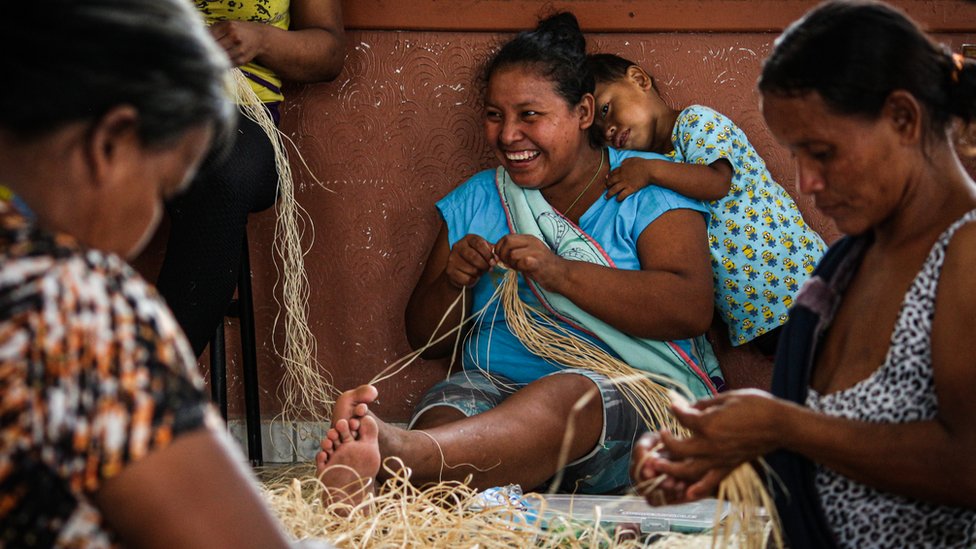 Três mulheres warao fazem artesanato, uma delas tem um grande sorriso e uma criança apoiada no ombro