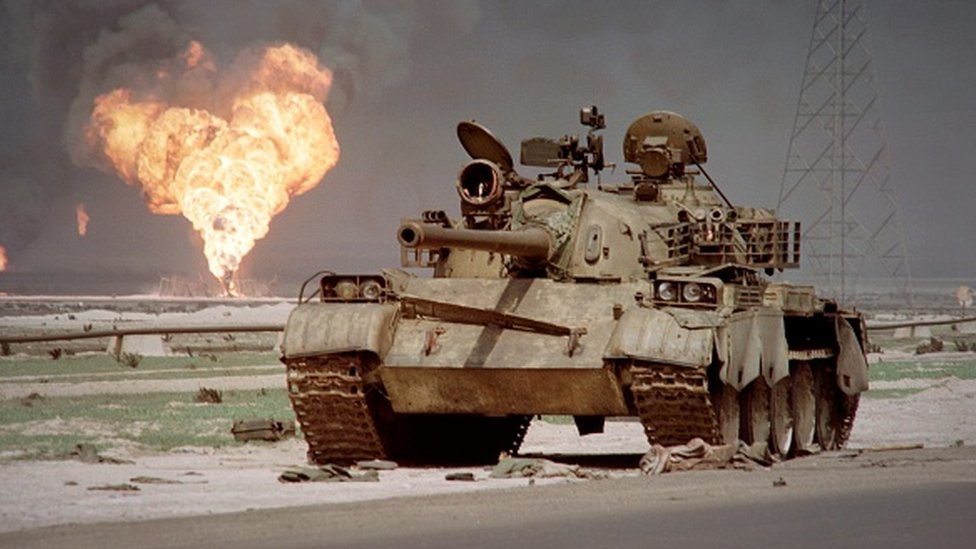 Un tanque abandonado en el desierto de Kuwait con un poso de petróleo en llamas en el fondo.