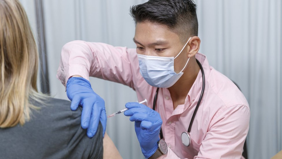 Un enfermero, con guantes y mascarilla, pone una vacuna en el brazo de una mujer.