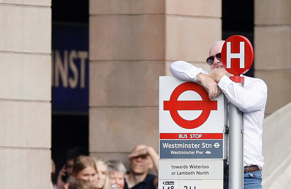 A man climbing a bus stop sign trying to see the coffin being carried into Westminster