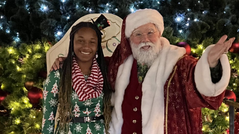 Un intérprete de Santa, junto a una intérprete de elfo, al frente de un árbol de Navidad