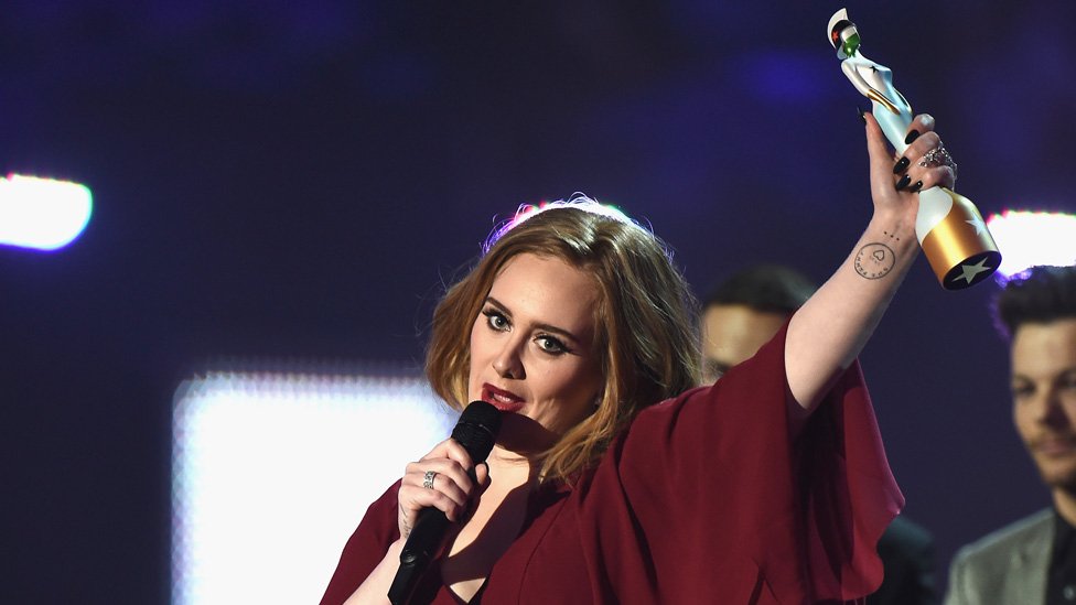 Em cerimônia, Adele segura microfone com uma mão e troféu em outra