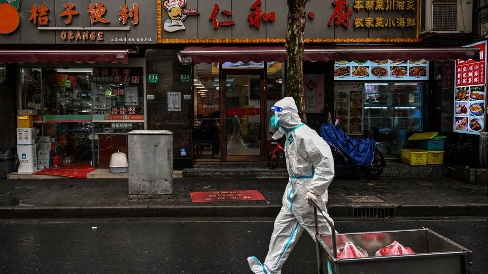 A worker covered from head to toe in a white protective suit pulls a cart with meat inside past restaurants
