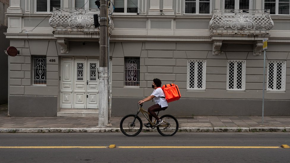 Franciele de bicicleta pedalando na rua, com mochila de delivery