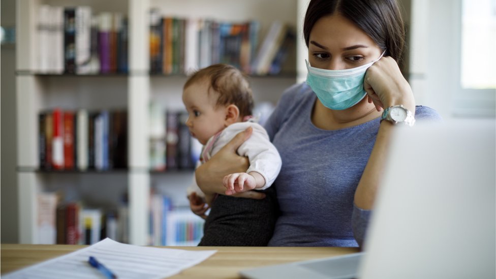 Mulher de máscara com bebê no colo, sentada em frente a mesa com laptop