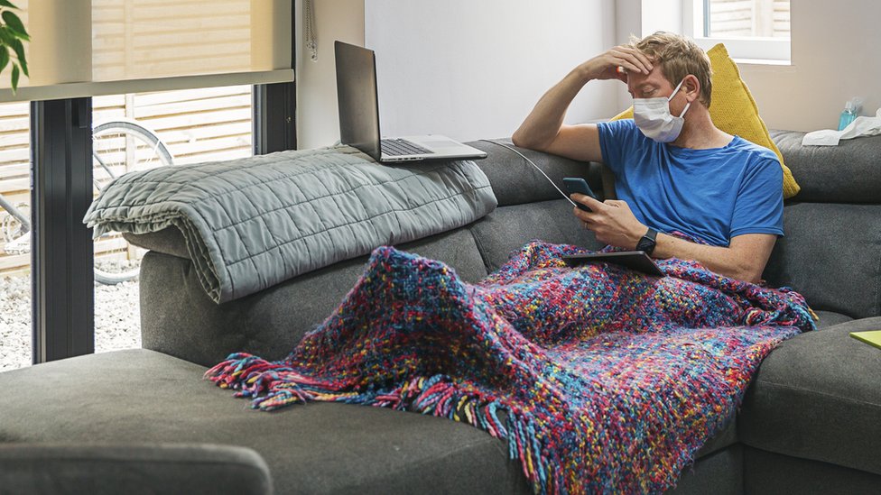Rapaz de máscara usando celular sentado no sofá com cobertor e mão na cabeça