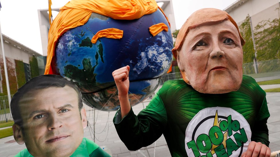 Два протестующих в масках канцлера Германии Ангелы Меркель (справа) и президента Франции Эммануэля Макрона (слева) проводят демонстрацию перед зданием канцелярии Германии перед прибытием европейских лидеров в Берлин, Германия, 29 июня 2017 г.
