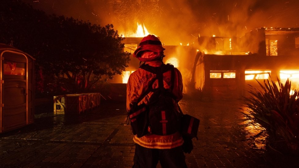 Bombeiro de costas trabalha de noite, diante de casas em chamas