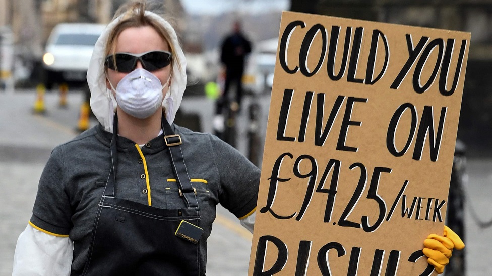 Женщина в защитной маске и перчатках держит плакат с жалобой на отсутствие поддержки для самозанятых и контрактных рабочих на Королевской Миле в Эдинбурге, Шотландия, 23 марта