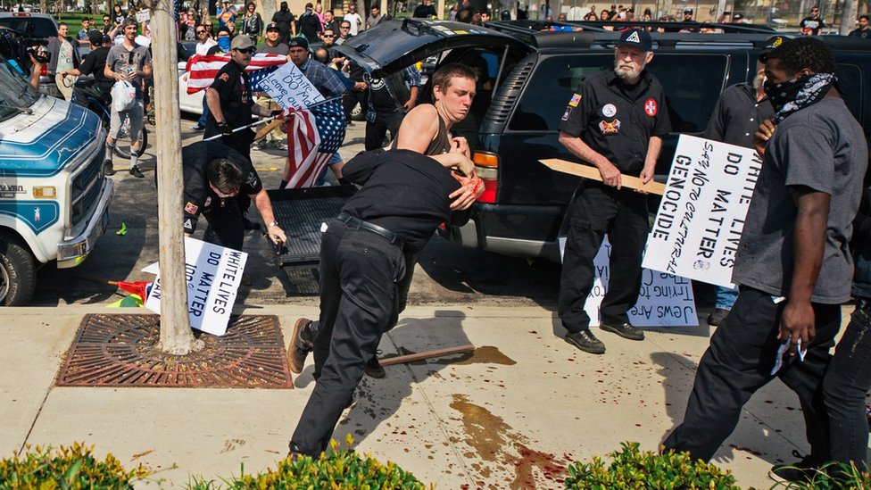 На этой фотографии, предоставленной OC Weekly, изображены столкновения контр-протестующих с членом KKK, когда он наносит удар атакующему протестующему (в центре), когда члены KKK пытаются начать антииммиграционный митинг в Пирсон-парке в Анахайме в субботу, 27 февраля 2016 г.