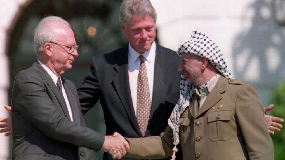 Билл Клинтон стоит между палестинским лидером Ясиром Арафатом, когда он пожимает руку премьер-министру Израиля Ицхаку Рабину после подписания мирного соглашения по Ближнему Востоку 13 сентября 1993 г.